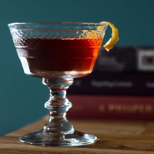 Reddish cocktail in glass with lemon peel garnish