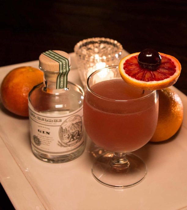 A small bottle of gin next to a big round cocktail in a glass with an orange wheel garnish